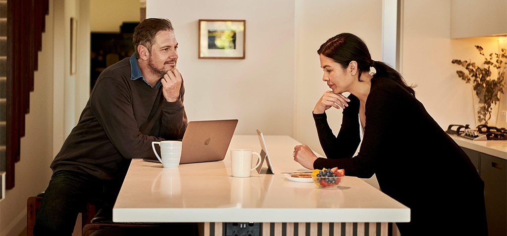 A man and woman sitting at a kitchen island, each using a laptop, with coffee mugs and a bowl of fruit on the counter.