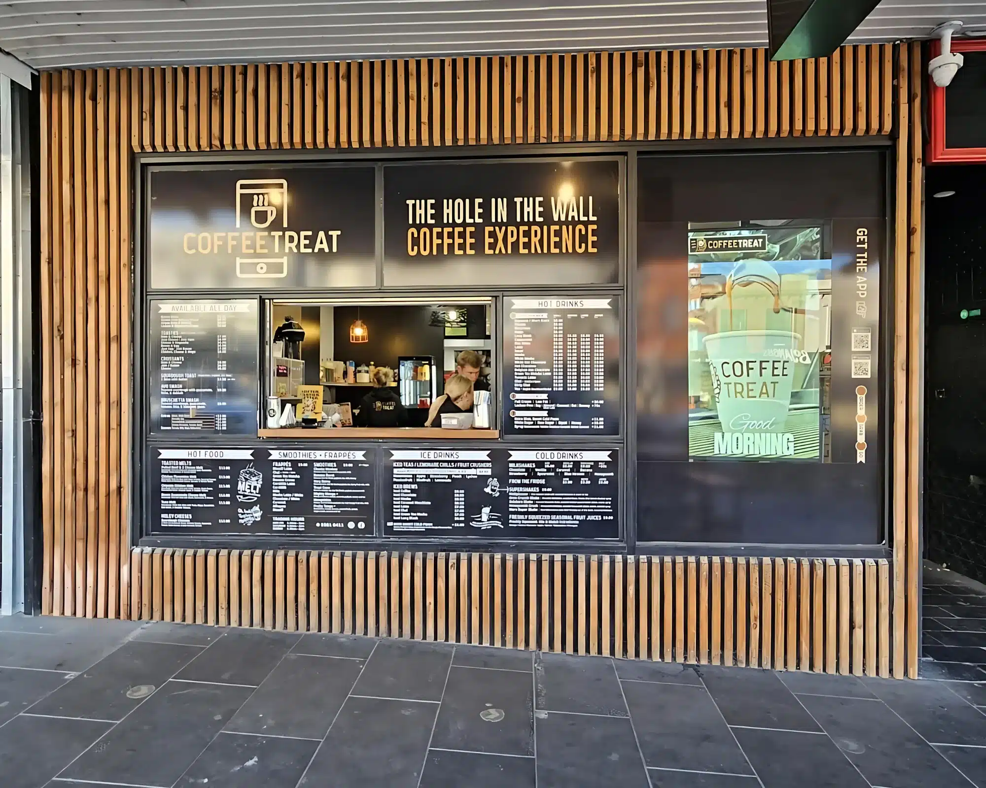 Street-facing coffee shop named "Coffee Treat" with a service window and a large black and wood-paneled exterior. A staff member is preparing drinks inside.