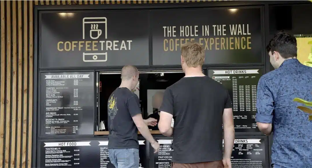 Three men ordering coffee at a walk-up coffee shop called CoffeeTreat, featuring a black storefront with menus and a sign that reads "The Hole in the Wall Coffee Experience."